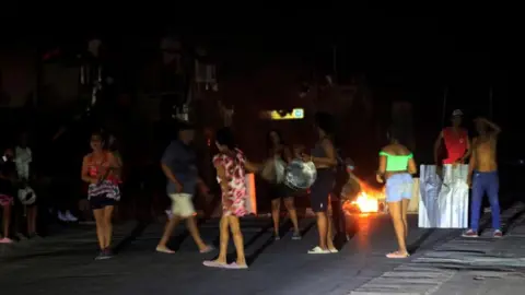 EPA/Shutterstock People protest against power outages in Havana on 7 July 2026. One person can be seen holding a large pot while another is banging a metal sheet. In the background burning rubbish can be seen on the street. 