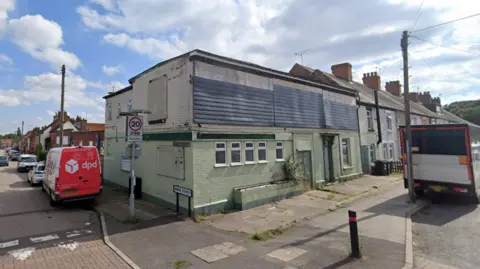 An old brick building painted green and white with metal sheets across the windows