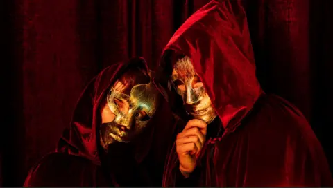 Conor Ross Photography Two people, pictured from the chest up, red cloaks with the hoods up. They are also wearing gold masks, covering their faces.