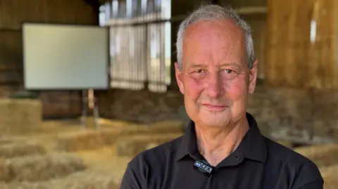Tim Dixon is stood in a shed with bales of straw around him. He has short grey hair and is wearing a black t-shirt. 