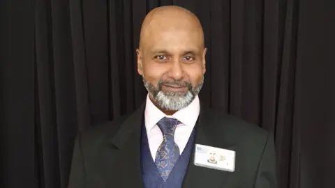 Supplied Mark Christopher, wearing a black suit jacket, mauve waistcoat, navy tie and white shirt. He is standing in front of black curtains and smiling at the camera.