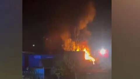 A screenshot of the fire filmed on a phone. The filming is across a road and there is a blue metal building in front of a blaze of fire at night time. 