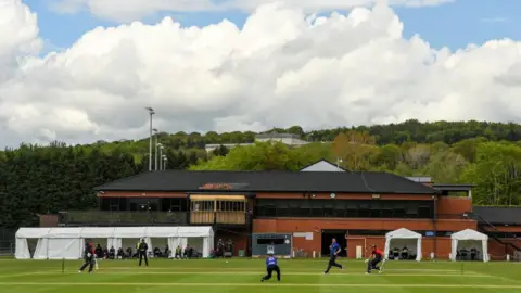 Getty Images A large red brick building with a black roof. In front of it, a green cricket pitch with several people running.
