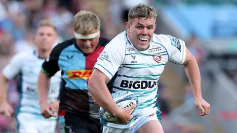 Jack Singleton runs with the ball tucked to his side held in his right hand during a match between Gloucester and Harlequins in May 2025. He is wearing a white Gloucester kit with blue horizontal stripes.