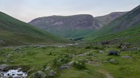 WMRT A grassy valley with slopes on either side. There is a river running through it with rocks all around. In the distance there is a large, imposing hill. The sky above is a blue-grey colour.