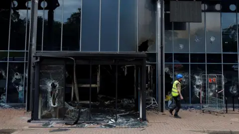 Reuters A close-up on the front entrance of a glass-fronted building - the entrance is seriously damaged, scorched from fire and with broken windows. The foyer entrance is strewn with fire debris