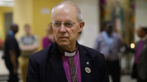 Reuters Archbishop of Canterbury Justin Welby looks on as he speaks with the press after a visit to the grave of Saint Oscar Arnulfo Romero, during a visit to El Salvador. Welby is wearing a black blazer with a bright purple shirt. A silver cross dangles from his neck against his chest.