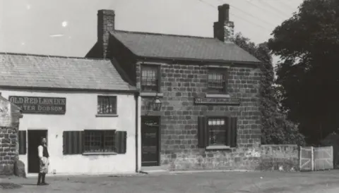 J W Hague A black and white photo of a brick building with a sign above the door reading "old red lion inn".