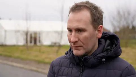 Rob Mathieson has short, light-brown hair and is wearing a navy-blue hooded puffer coat while standing in front of a grass field. Behind him, on the left-hand side, is a large white marquee tent. 