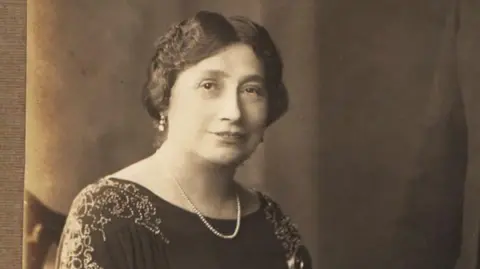 Rio Cinema Black and white photo of a woman wearing a necklace and earrings. She has short hair.
