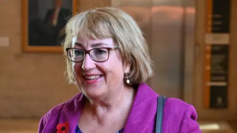 SNP MSP Annabelle Ewing on the way to First Minister's Questions in the Scottish Parliament. She has a short blonde bob haircut with a fringe and wears dark brown glasses. She also wears a purple blazer with a red and black poppy badge.