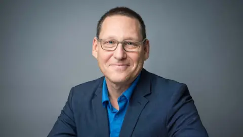 Avi Lewis wears a blue suit coat and blue collared shirt, no tie, and grey glasses as he smiles. Behind him is a neutral grey background 