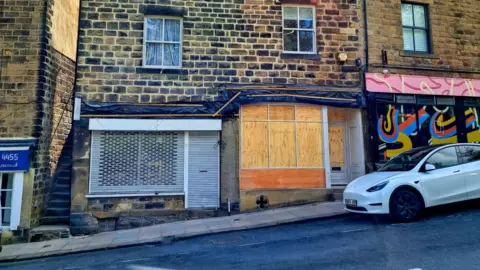 Leeds City Council A row of older stone buildings along a street, with the central building being the main focus. This building has three stories and is constructed from dark, weathered stone blocks. It features four windows on the upper floors, each with white frames and curtains visible inside. The ground floor has two sections: one on the left with a metal security shutter pulled down, and another on the right with a large wooden board covering the window and door.
To the right of the building, there is a neighbouring property with a colourful mural painted on its shopfront. A white car is parked on the street in front of the buildings.