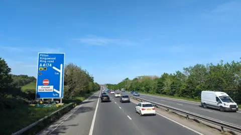 The M32 with vehicles travelling down carriageway and green field with phone tower in background. 