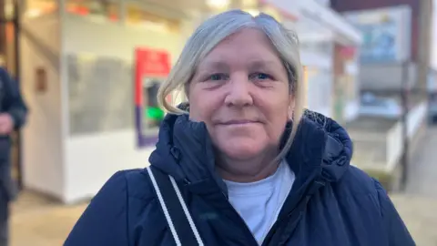 Ms Ross has her dyed blond hair tied back. She is wearing a white top and blue coat which is zipped up. The post office can be seen behind her.