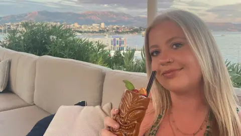 Amelia Sewell Amelia Sewell, 25 anos, cabelos loiros lisos e olhos azuis. Ele segura um copo tiki marrom perto do rosto com um canudo preto. Ela está sentada em um sofá creme e atrás dela você pode ver as montanhas. Um mar o separa dele e de um corpo de mar. 