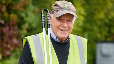 A man in a grey baseball cap and yellow high vis jacket smiles at the camera while holding a litter grabber