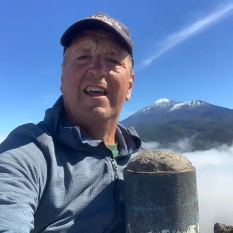David McNicol A man in a baseball cap with a blue top on standing at the top of a mountain or hill