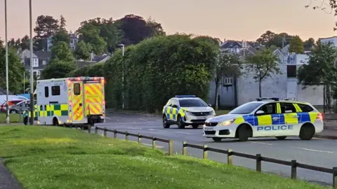 BBC Two police cars blocking a road, with an ambulance in the background
