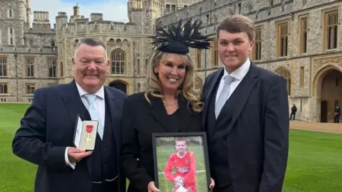 A middle-aged couple and a younger man - all wearing smart dark-coloured suits - pose for a photograph at Windsor Castle. The older man is holding an OBE and the woman is holding a framed photo of a boy dressed in a red football kit. 