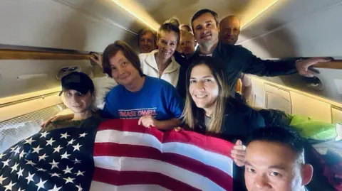 Truthsocial.com/realDonaldTrump Gilman, în stânga, văzut cu un steag american arborat peste el în avion. El este lângă alte câteva persoane, inclusiv mama sa.