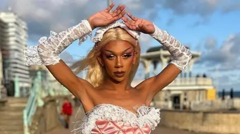 Raiine Kane, as Raiine, pictured outside on Brighton sea front. Raiine wears a pale pink bustier top with a white ostrich feather trim and matching white lace gloves. She wears a long blonde wig and raises her arms above her head. 