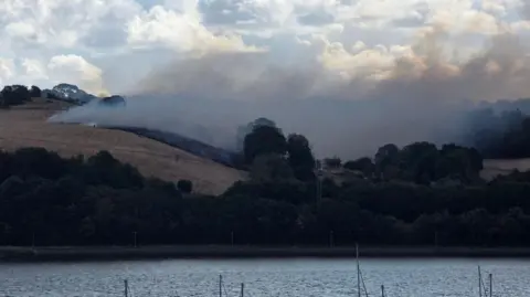 Tim Lee A wide view of the area showing a rolling hill on fire with lots of smoke. 