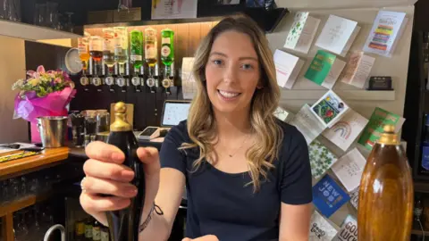 A young woman with long blonde hair standing behind a bar. She has her right hand on a beer pump and is smiling at the camera. There is a row of optics in the background and some well done cards on a board.
