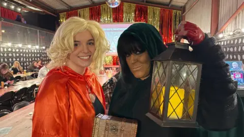 Two women in fancy dress, one in a blonde wig and a red cloak, and the other in a dark cloak, fake tan and heavy eye-liner, while holding a lantern, pose for a picture in a large bar area