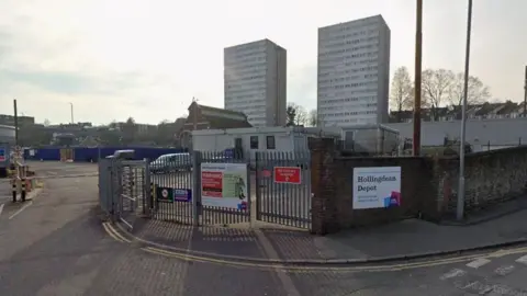 A Google StreetView image showing the entrance to the Hollingdean Depot which has large metal gates and fences and a council sign on the front with the name of the depot on it. In the background are two tower blocks.