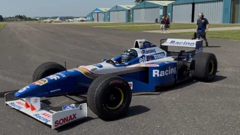 1996 F1 Williams car - which is mostly blue with some white on the body and blue, white and yellow branding, on the runway at Cotswold Airport on a sunny day. There are blue hangars and other planes on the edge of the airfield and a few members of staff are stood behind the car.