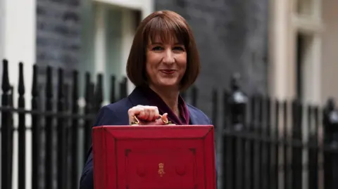 PA Media Rachel Reeves dressed in a blue suit and a maroon scarf holding a red briefcase. Behind her are black metal railings and behind them the blurred sides of buildings.