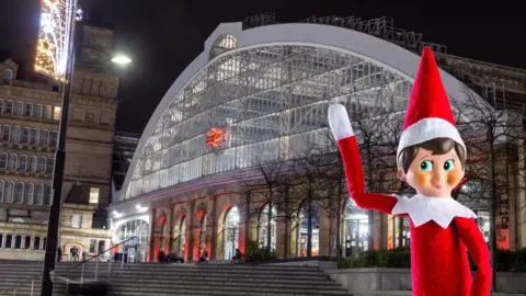 Liverpool BID Liverpool Lime Street train station entrance sits on top of steps, its lit up as it is night time and there's a black lamp post to the left of the picture which has a lighted Christmas decoration. To the right of the picture is a super imposed toy elf waving