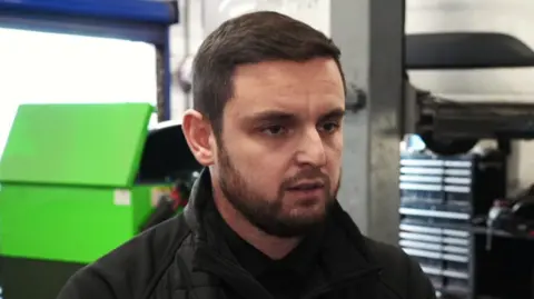 A man with short brown hair and a short beard looks away from the camera. He is wearing a black jacket and standing in a mechanic's workshop