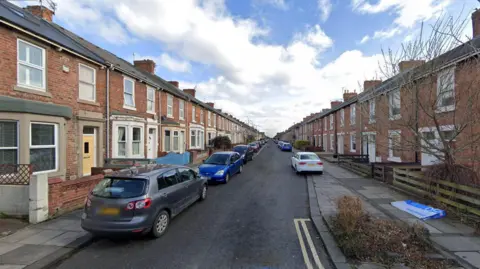 Bolingbroke Street is lined with two-storey red-brick terraced houses. Cars are parked all the way along.