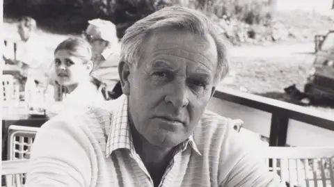 A black and white photo of a man wearing a long-sleeved pale top with the sleeves rolled up. He's sitting at a table and leaning forward on his elbows, and looking, unsmiling, at the camera. There's a wooden railing and some other people sitting at tables behind him.