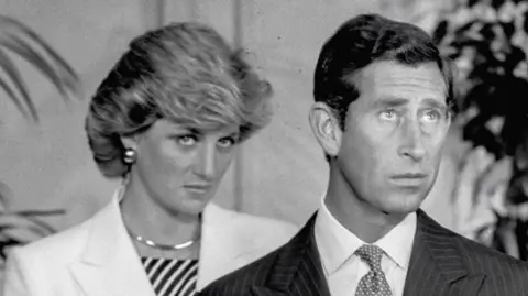 Richard Blanshard A black and white image of Charles and Diana who are both looking up and look unhappy. Diana is standing behind Charles and wearing a white jacket and striped top with large earrings. Charles is wearing a pinstriped suit and spotty tie. 