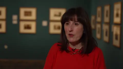 Imogen Holmes-Roe stands in front of the Turner collection. She has shoulder length dark hair, wears a red top and a chunky, gold necklace.