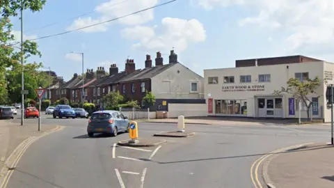 The junction of Ber Street and Queens Road. A blue car is waiting at the junction for another blue car to pass. A white building is on the opposite side of the road and to the left of it is red-brick terraced houses.