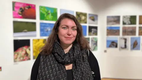 Louise Le Pelley is smiling with green eyes and brown hair worn loosely. She is wearing a black cardigan and patterned scarf. In the blurred background is wildlife photos in an exhibition. 