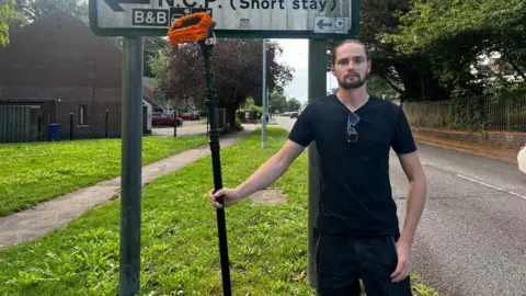 Lily-May Symonds/BBC Liam Wildish is standing in front of a road sign holding a large brush. He's dressed in a black t-shirt and cargo trousers. He has a short beard and long hair tied back.