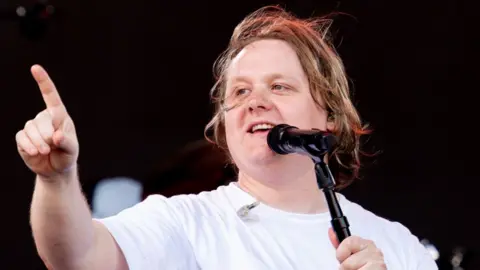 Lewis Capaldi on stage at Glastonbury 2023 in a white T-shirt singing into a microphone with one finger pointing in the air