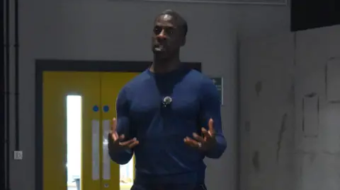 Eastern Education Group Dwain Chambers wearing a navy jumper and standing up while seemingly addressing a group of people who are out of shot. He is making gestures with his hands.