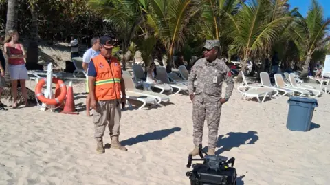 Oficiais da Defensa Civil Dominicana procuram Sudiksha Konanki. Eles estão cercados por areia e por cadeiras de praia vazias e um drone está na areia perto de seus pés 