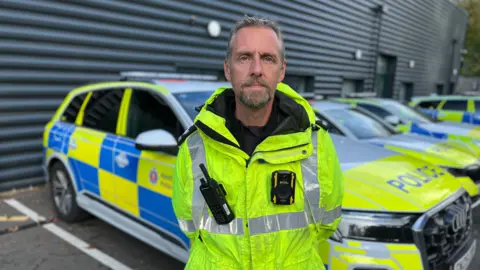 Chris Wade is wearing a police uniform and a high vis coat and he's standing in front of a row of police cars at a Kent Police compound