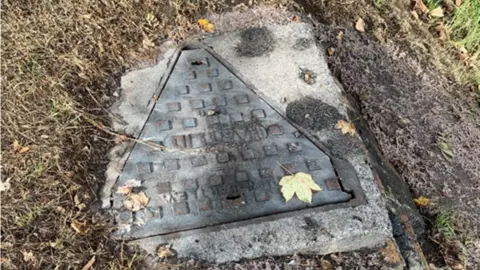 Wessex Water The manhole cover for the sewer that overflowed. The cover is a triangle shape and has a square of concrete around it. There is grass around the concrete.