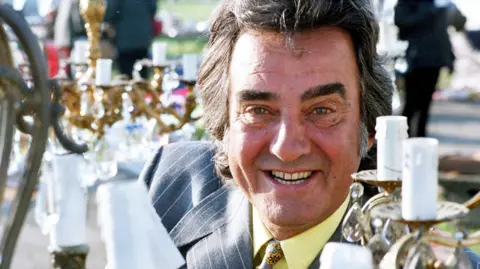 Antiques expert and TV personality David Dickinson with his famous tanned skin wearing a yellow shirt and grey suit with blue pinstripes. He's pictured among several candelabras at an antiques fair. 