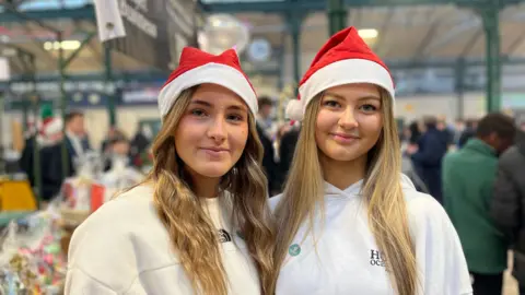 Two girls with red Santa hats. They are wearing white jumpers and smiling. Both have long blonde hair and are standing in the middle of St George's Market.