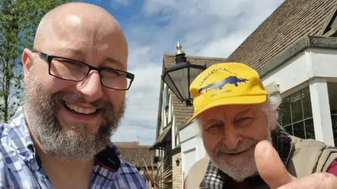 Richard Mourton A selfie of Richard, a man, with long white beard. His dad, a man in a yellow hat, who also has a long white beard, is standing next to him. They are smiling for the camera.