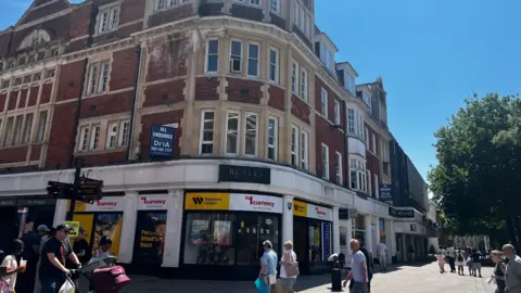 Alex Pope/BBC A historic red-brick corner building housing retail units on a busy pedestrian shopping street under clear blue skies.